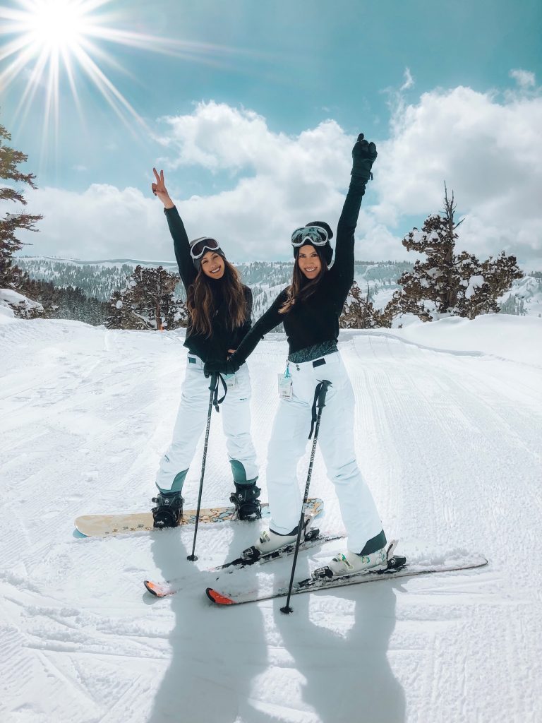 Deux jeunes filles sur leurs skis, le bras en l'air et le sourire aux lèvres, en train de poser pour la photo avec un grand soleil en fond.